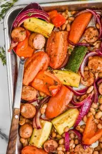 overhead view of a spoon in a pan of sausage, beans and roasted vegetables to make a sheet pan dinner