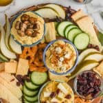 overhead view of a hummus charcuterie board with three different topped hummus bowls, cucumber slices, pieces of cheese, slices of pear, pretzel flats, and cranberries