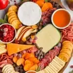 overhead view of an oval wood board with four varieties of cheese cut and whole, sliced pears, three small bowls of spreads, pear slices, persimmon slices, and hard meats