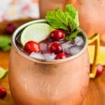 cranberry moscow mule in a copper mug garnished with fresh cranberries, lime wedges, and mint springs on a wooden tray