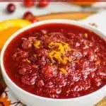 cranberry sauce in a white bowl with orange zest on top, sitting on a pieces of burlap and a fall napkin with cinnamon sticks and fresh cranberries around