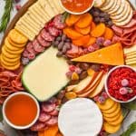 overhead view of an oval wood board with four varieties of cheese cut and whole, sliced pears, three small bowls of spreads, pear slices, persimmon slices, and hard meats