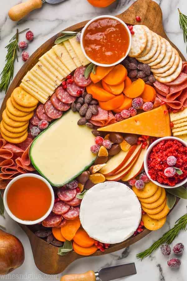 overhead view of an oval wood board with four varieties of cheese cut and whole, sliced pears, three small bowls of spreads, pear slices, persimmon slices, and hard meats