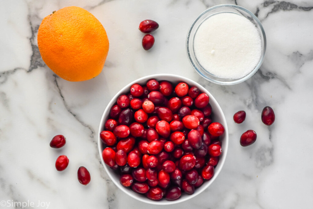 overhead photo of a bowl of fresh cranberries, an orange, and a bowl of sugar