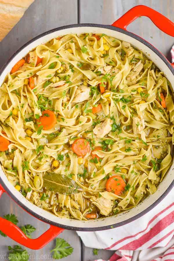 overhead view of a red handled stock pot full of turkey noodle soup - long noodles, pieces of turkey, a bay leaf, carrots, and corn are visible