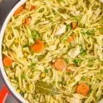 overhead view of a red handled stock pot full of turkey noodle soup - long noodles, pieces of turkey, a bay leaf, carrots, and corn are visible