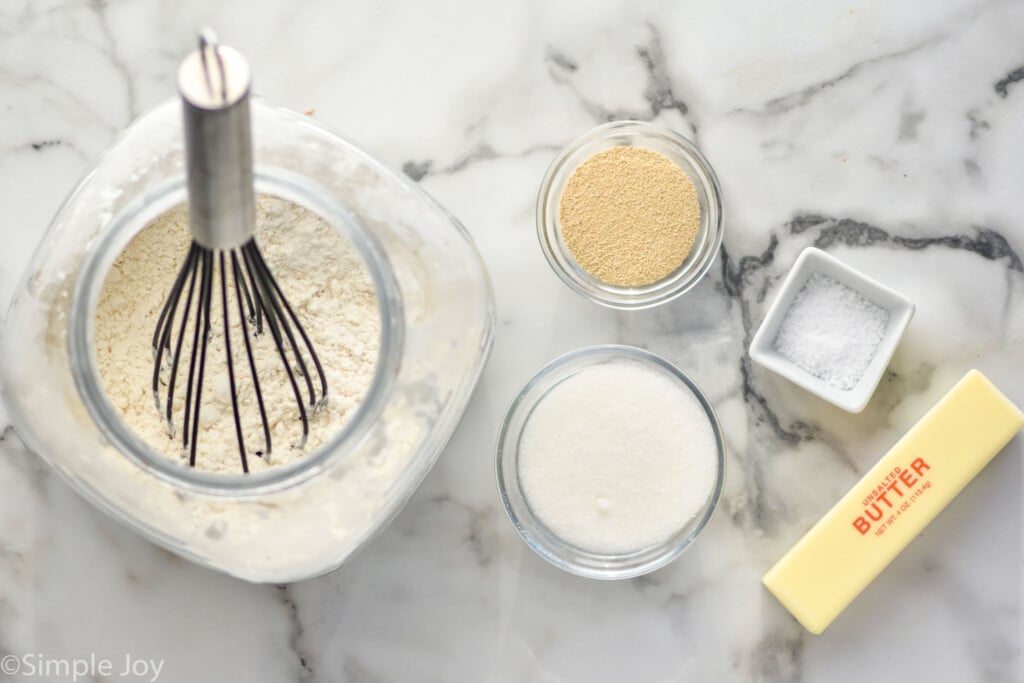 overhead photo of a jar with flour, a bowl with sugar, a bowl with yeast, a bowl with kosher salt, and a stick of butter