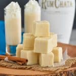 a pile of white rum Chata fudge on a wood board, with cinnamon sticks next to it, two tall shot glasses full of rum Chata in the background and a bottle of rum Chata in the distance