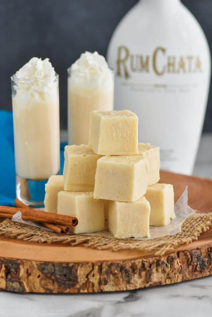 a pile of white rum Chata fudge on a wood board, with cinnamon sticks next to it, two tall shot glasses full of rum Chata in the background and a bottle of rum Chata in the distance