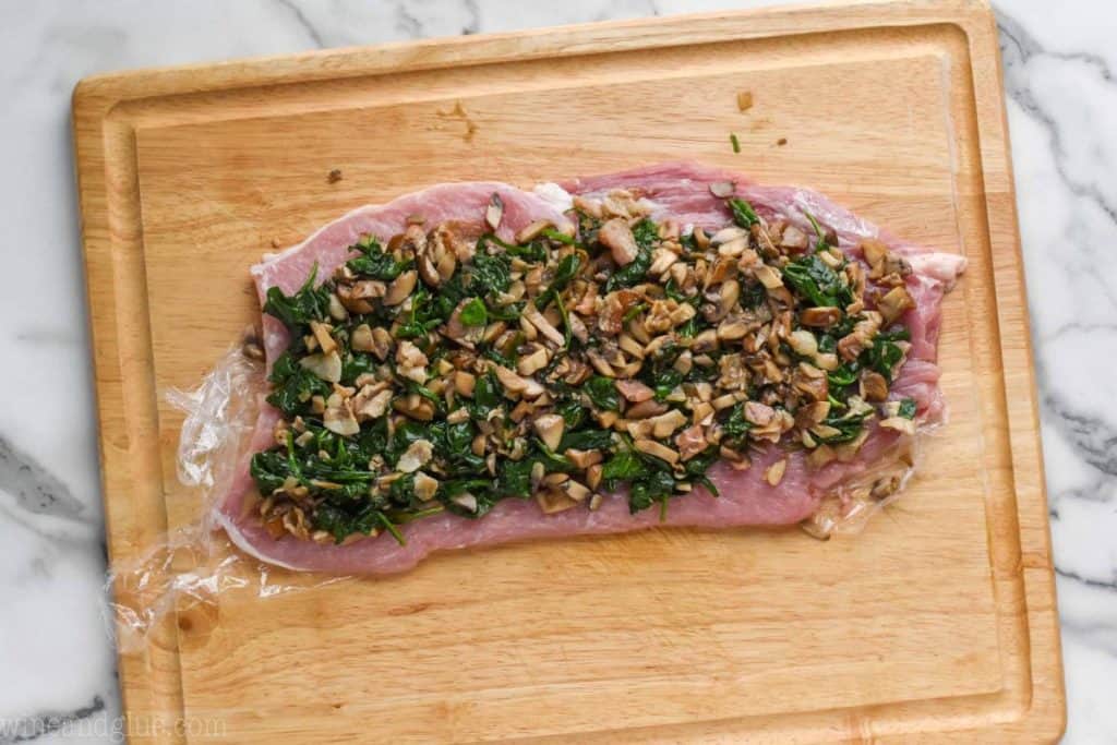 a flattened pork tenderloin on a cutting board that is covered in spinach and mushrooms filling