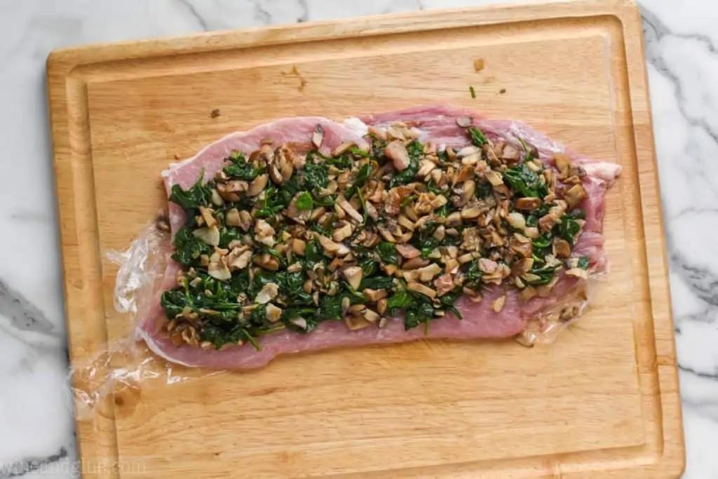 a flattened pork tenderloin on a cutting board that is covered in spinach and mushrooms filling
