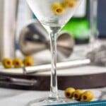 pinterest graphic of a gin martini in a chilled glass with three olives on a spear and bottles in the background, says: "the perfect gin martini, simplejoy.com"