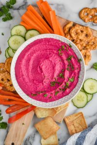 overhead view of beet hummus recipe in a white bowl in a cutting board surrounded by cut carrots, pretzel flats, cucumber slices, red pepper slices, and pita chips