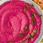 overhead view of a white bowl full of bright pink beet hummus garnished with fresh parsley