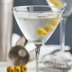 a frosted martini glass that has been chilled full of a gin martini recipe with three olives on a toothpick, a metal jigger and another gin martini on a tray in the background