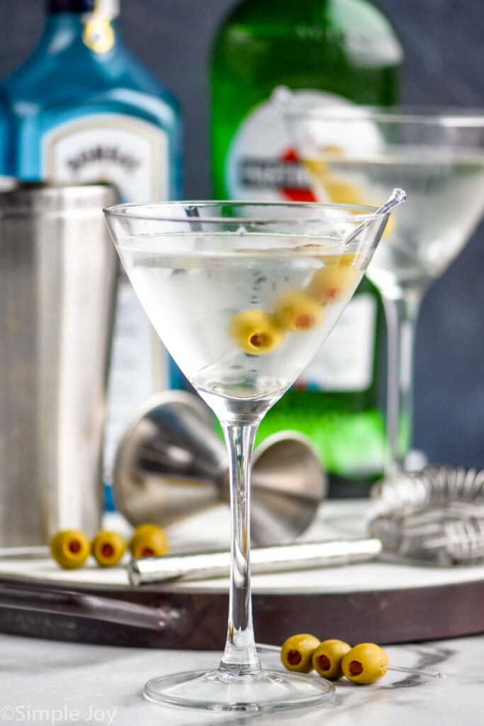 a gin martini in a frosted martini glass with a tray full of bar tools, a bottle of Bombay saphhire, and a bottle of dry vermouth in the background