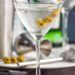 a gin martini in a chilled glass with three olives on a spear and bottles in the background