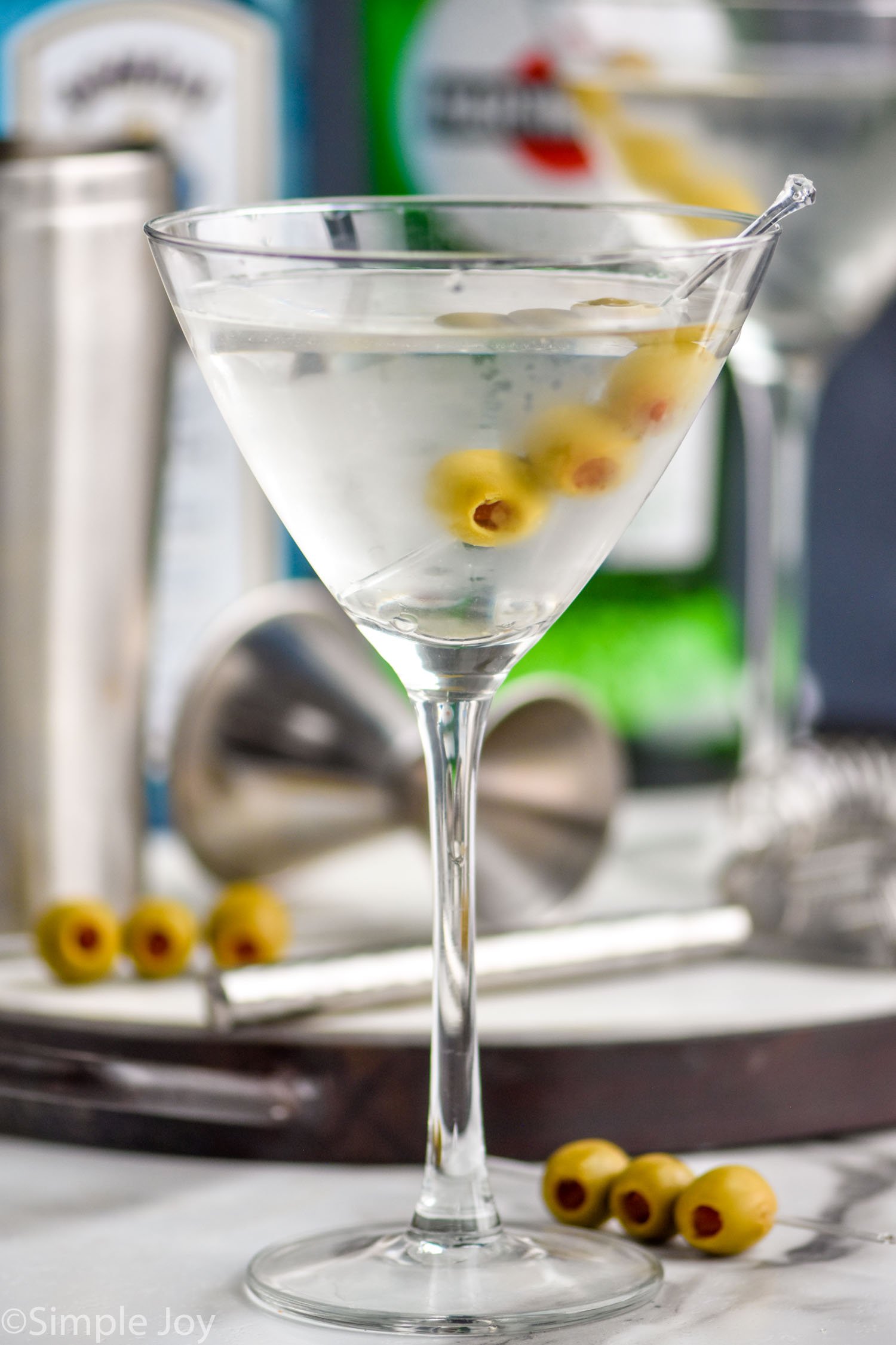 a gin martini in a chilled glass with three olives on a spear and bottles in the background