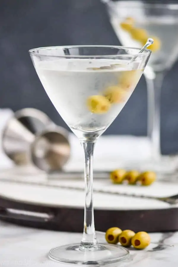 a frosted martini glass that has been chilled full of a gin martini recipe with three olives on a toothpick, a metal jigger and another gin martini on a tray in the background