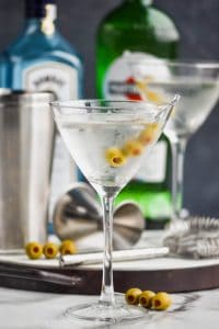 a gin martini in a frosted martini glass with a tray full of bar tools, a bottle of Bombay saphhire, and a bottle of dry vermouth in the background