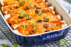 side view of a blue ceramic baking dish filled with ground beef enchiladas sitting on a wire cooling rack with cilantro around it