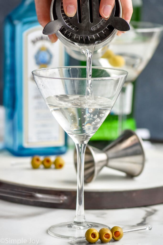 straining a gin martini into a martini glass with a bottle of Bombay sapphire on a tray in the background and a metal jigger on the tray