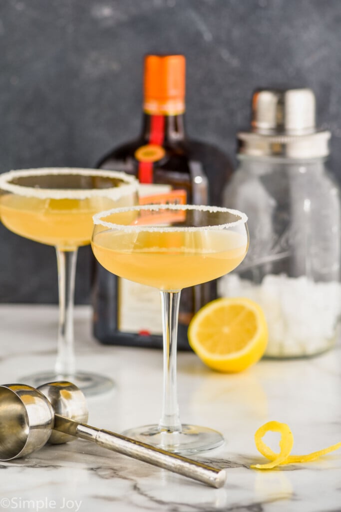 two coupe glasses rimmed with sugar holding a sidecar recipe with a double sided metal jigger in the foreground, a lemon twist off to the side and a bottle of cointreau and a cocktail shaker in the background