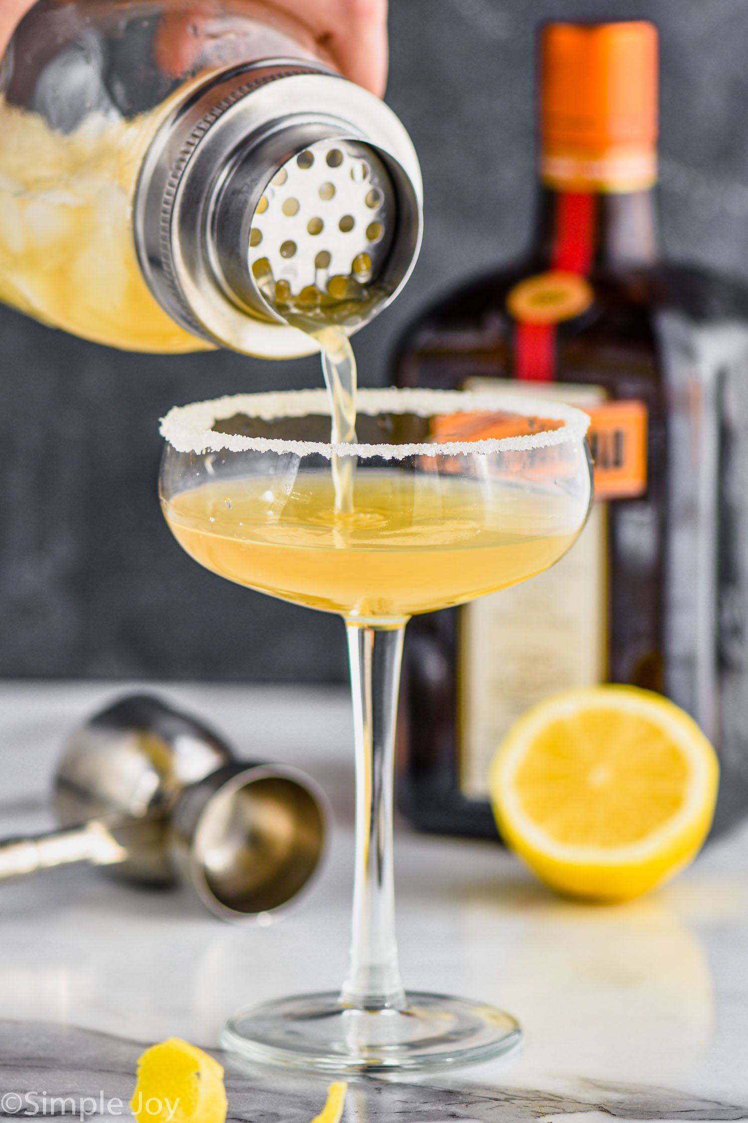 a cocktail shaker pouring a sidecar cocktail into a coupe glass rimmed with sugar