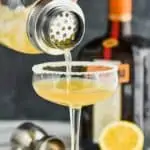 a cocktail shaker pouring a sidecar cocktail into a coupe glass rimmed with sugar