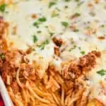 a close up of a white ceramic baking dish full of baked spaghetti topped with cheese and fresh parsley