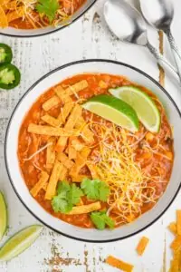 overhead view of enchilada soup in a white bowl garnished with chip strips, lime wedges, cilantro, and shredded cheese
