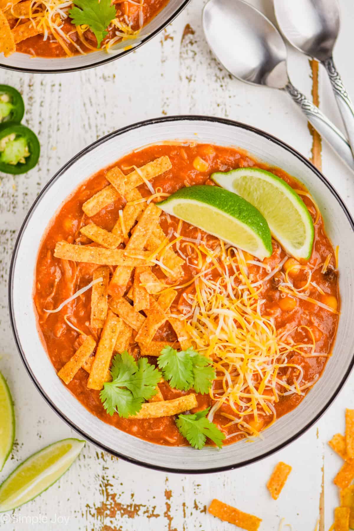 overhead view of enchilada soup in a white bowl garnished with chip strips, lime wedges, cilantro, and shredded cheese