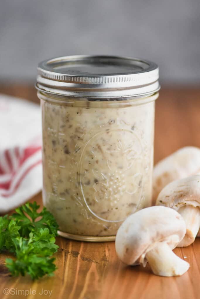 a mason jar containing condensed cream of mushroom soup with three fresh mushrooms sitting on one side of it and fresh parsley on the other, a cloth napkin in the background
