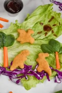 a piece of iceberg lettuce serves as the background for a dinosaur scene made out of veggies and dinosaur chicken nuggets