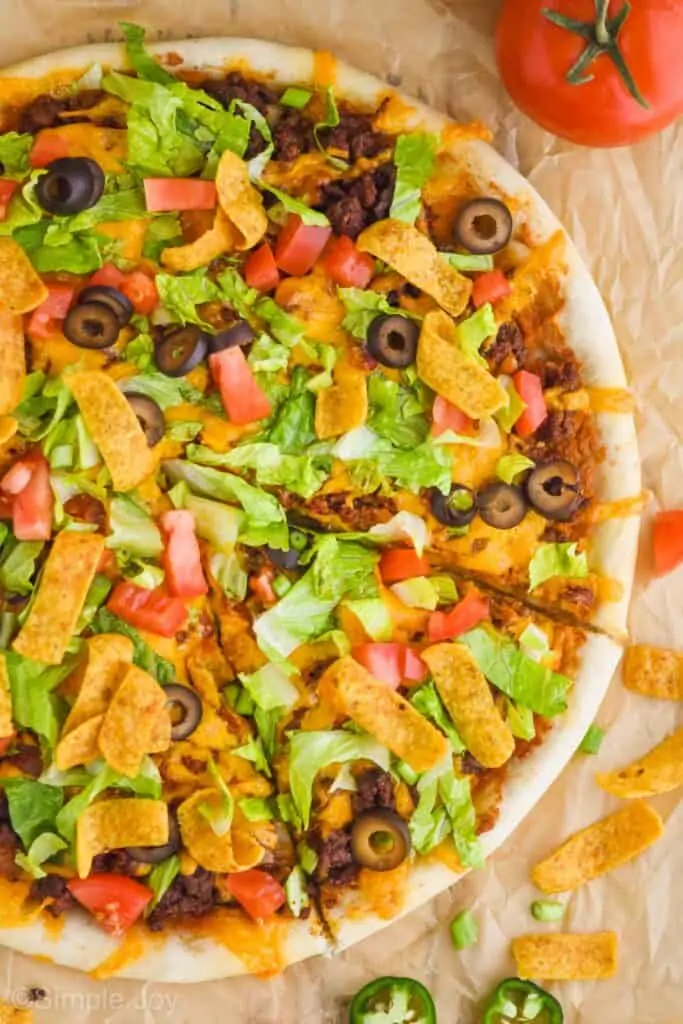 overhead photo of a taco pizza that has been topped with olives, lettuce, diced tomatoes, and green onions with one slice cut out