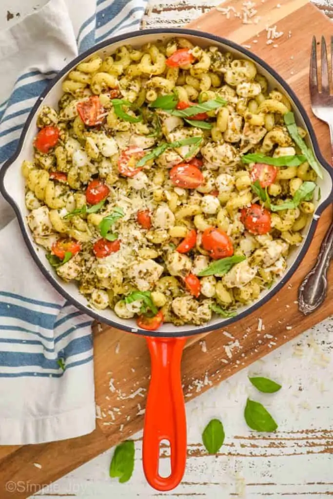 pulled back overhead view of a red handled skillet with cavatapi noodles, pesto sauce, tomatoes, and small mozzarella pearls on a cutting board with a cloth napkin