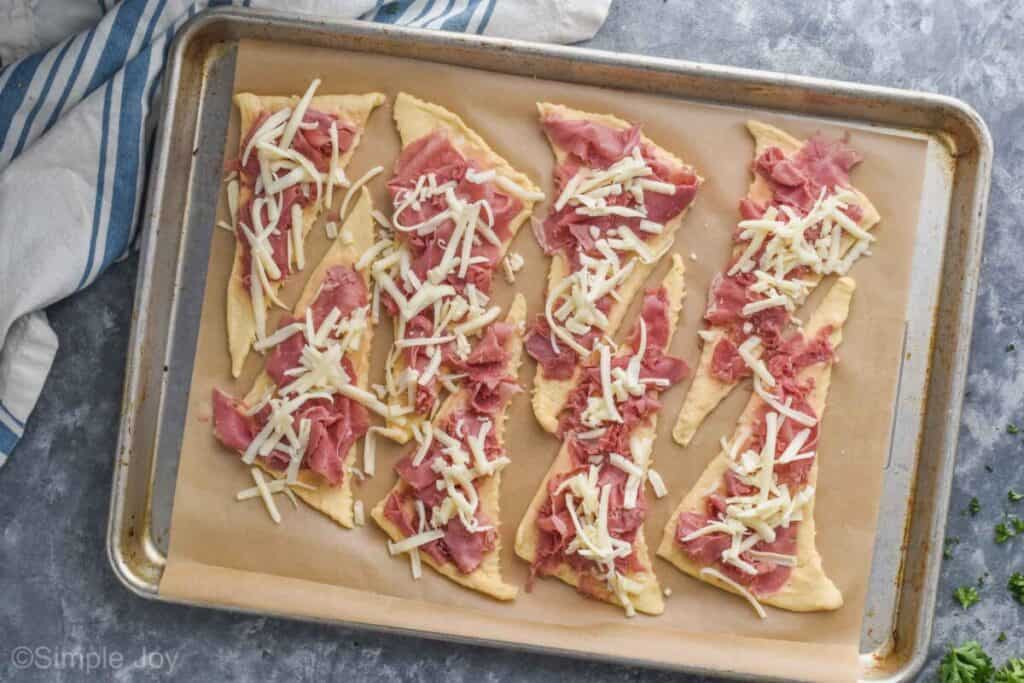 overhead of a baking sheet with 8 unrolled crescent rolls that have corned beef and shredded Swiss cheese on them