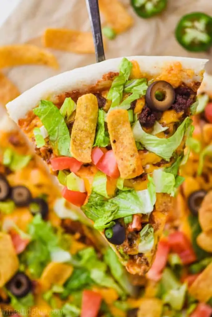 a slice of taco pizza that is being held up high over the rest of the pizza