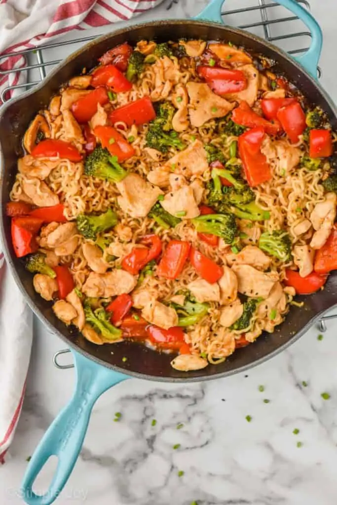 overhead view of a teal skillet on a wire rack on a marble counter full of stir fry noodles with chicken, ramen, red peppers and broccoli