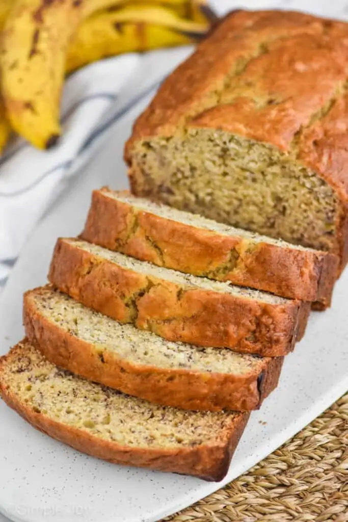 side view of best banana bread recipe where the first half of the loaf has been sliced, ripe bananas in the background