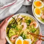 overhead view of two hands holding a bowl of easy ramen with a soft boiled egg, chopsticks, shrimp, and broccoli