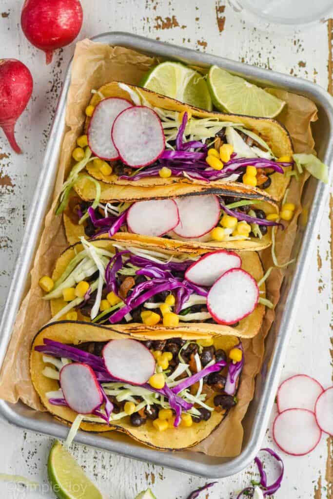 overhead of four vegan black bean tacos on a rimmed baking tray, garnished with sliced radishes, shredded cabbage, and corn