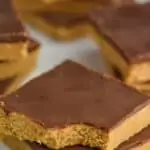 two square chocolate peanut butters, one with a bite out of it stacked on another on a white surface, more in the background