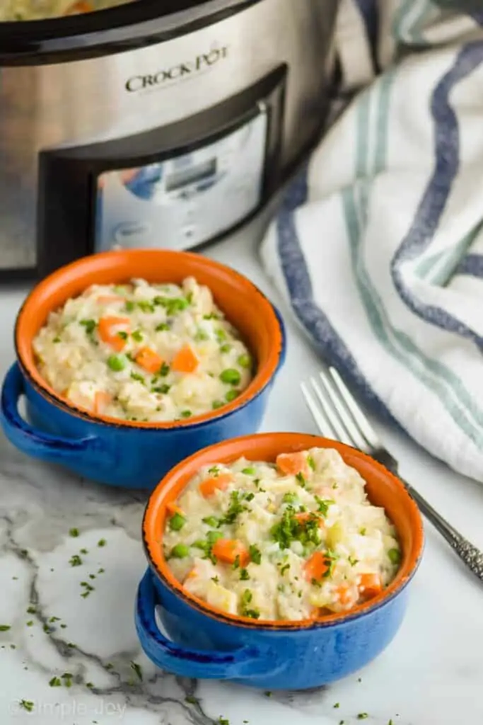 two small blow soup bowls holding creamy chicken and rice casserole on a marble countertop with a crockpot behind them