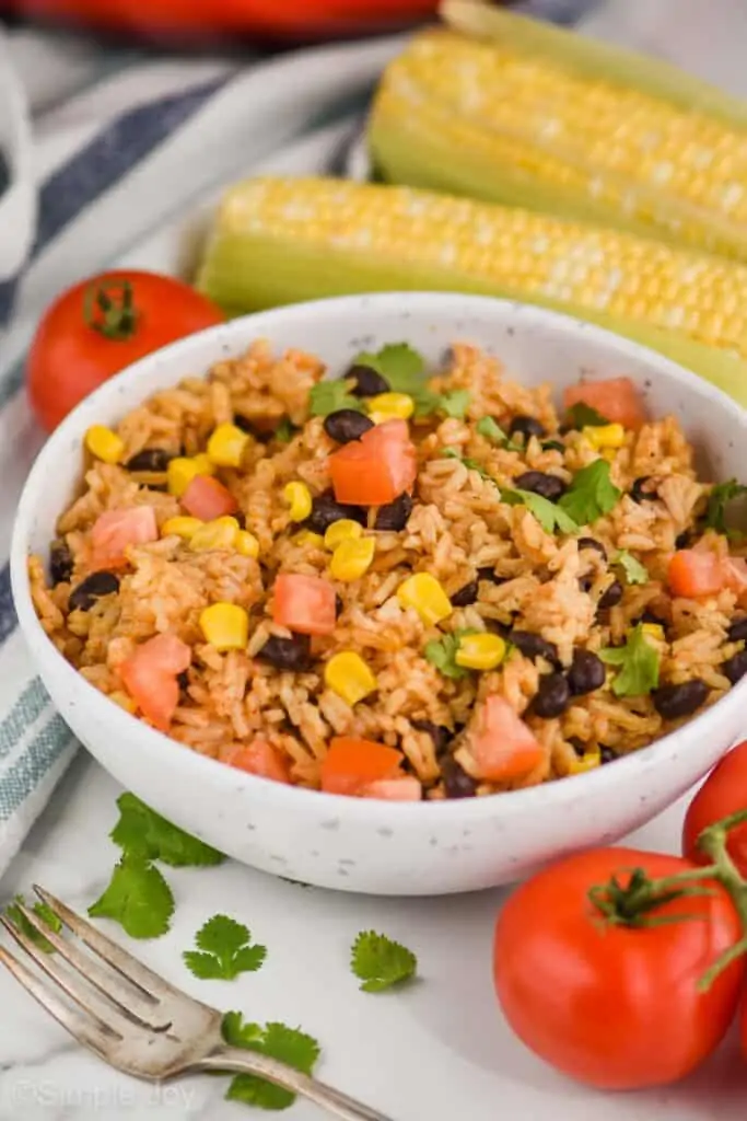 side view of a speckled white bowl with easy black beans and rice recipe with corn, tomatoes, and cilantro, fresh corn and tomatoes in the background