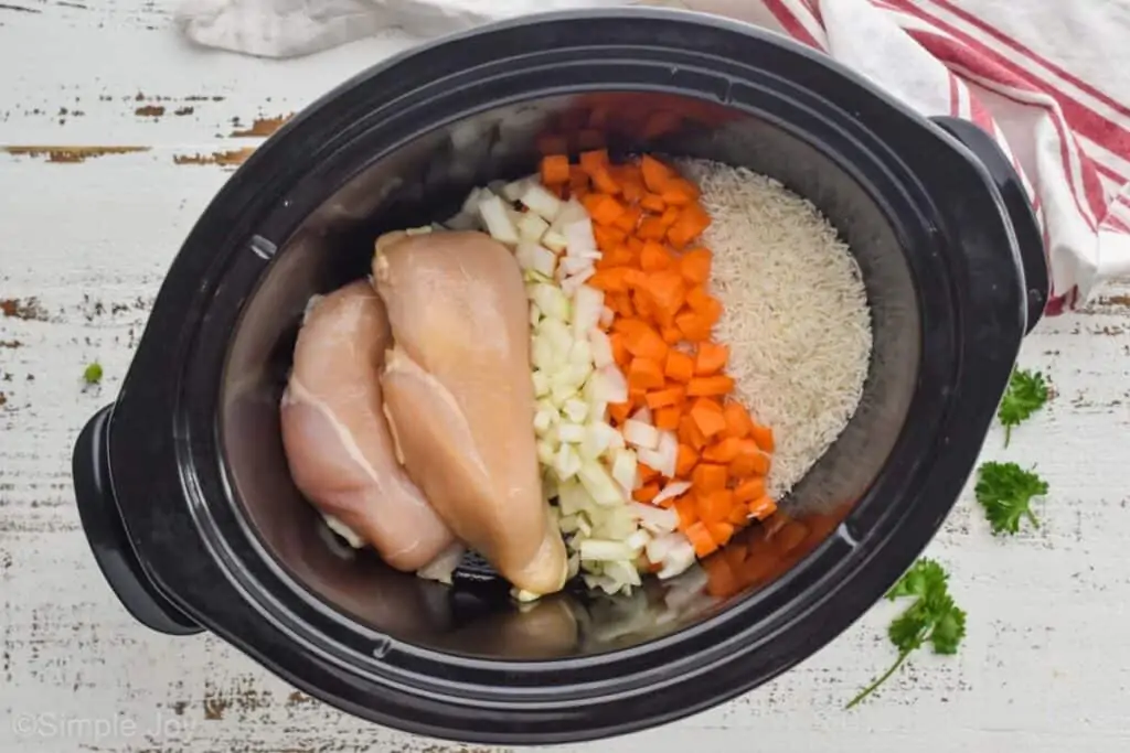 overhead of a slow cooker with raw chicken, onion, carrot and uncooked rice ready to make chicken and rice casserole