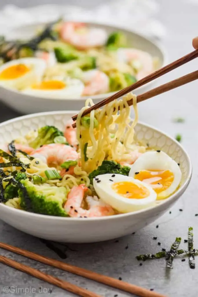 chopsticks lifting ramen noodles out of a bowl that also has broccoli, shrimp, and a soft boiled egg