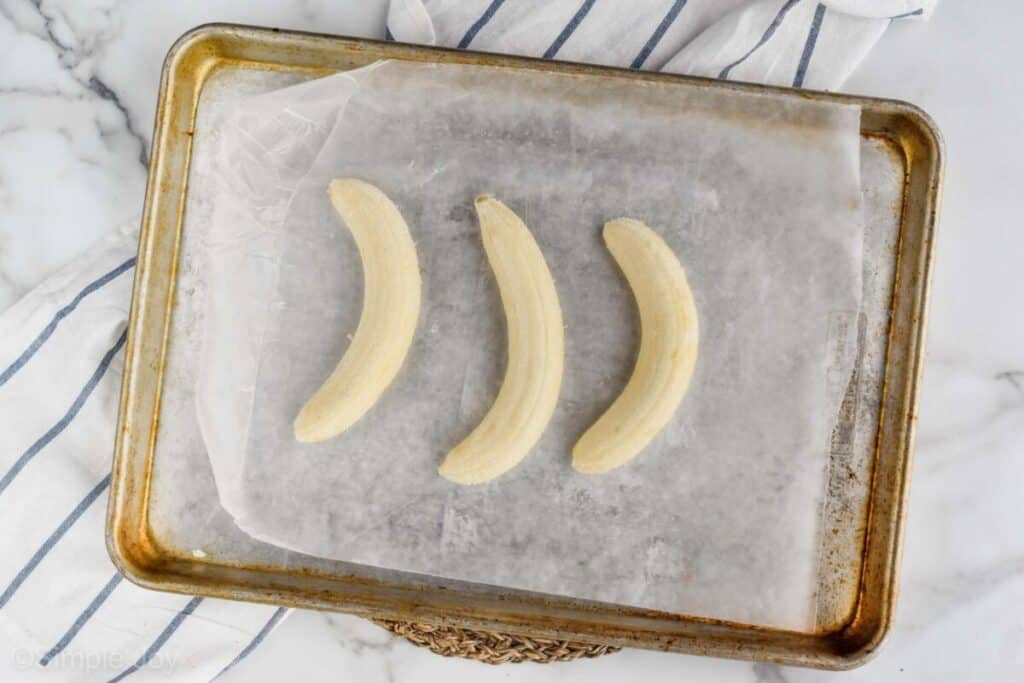 overhead view of three bananas that have been peeled and are sitting on a wax paper lined baking sheet to be frozen