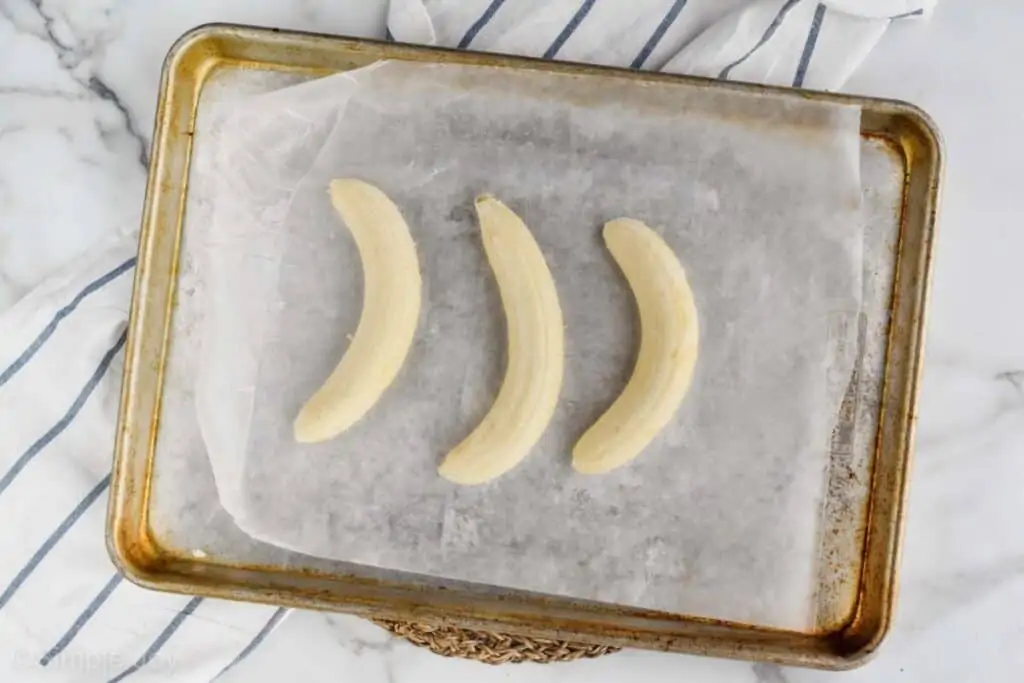 overhead view of three bananas that have been peeled and are sitting on a wax paper lined baking sheet to be frozen