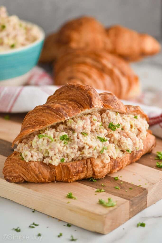 a ham salad sandwich made with a croissant on a cutting board with more croissants in the background and a bowl filled with ham salad